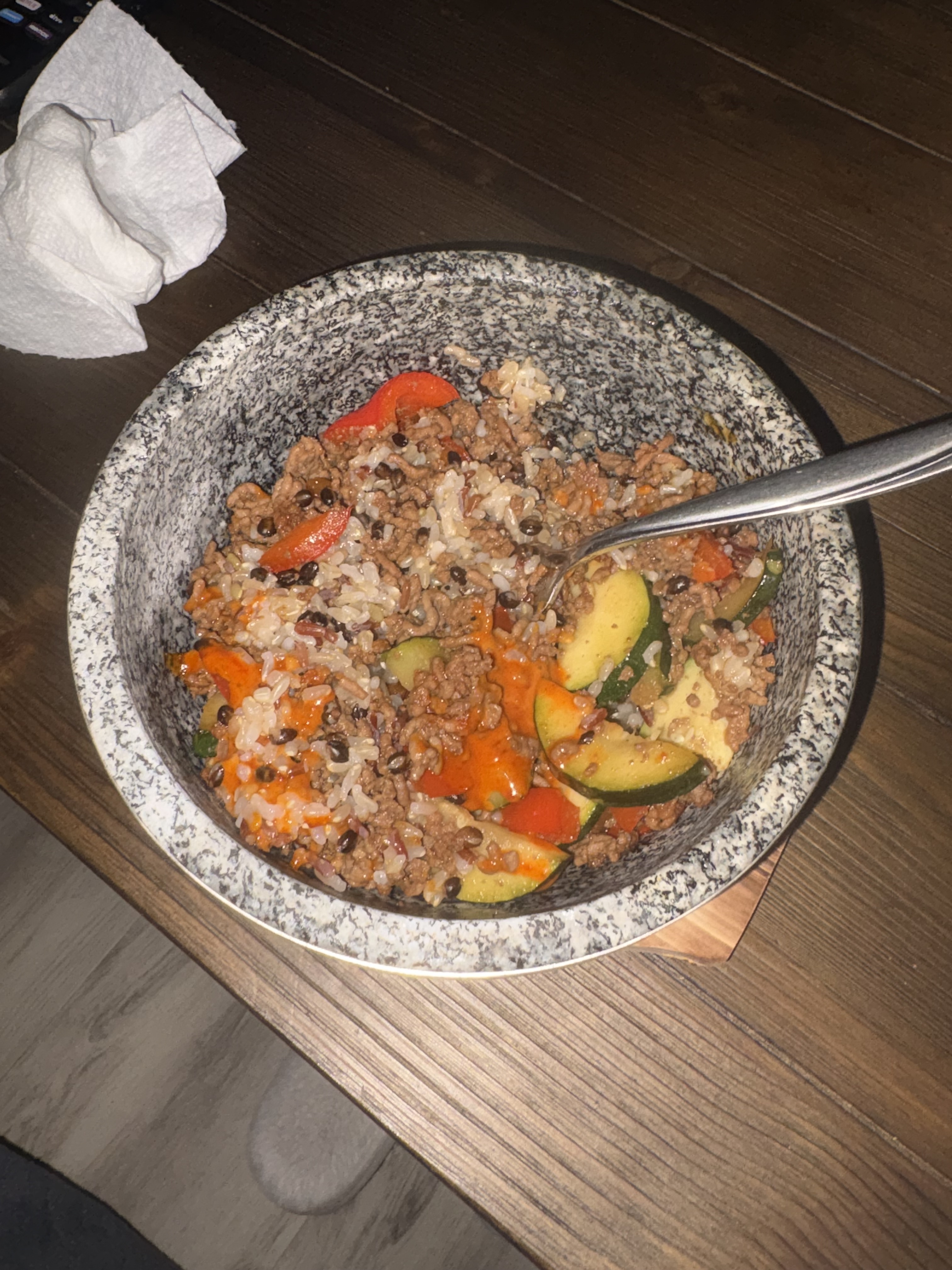 A speckled stone bowl on a wooden base holds rice, ground beef, zucchini, red bell pepper and orange sauce, with a spoon resting inside.