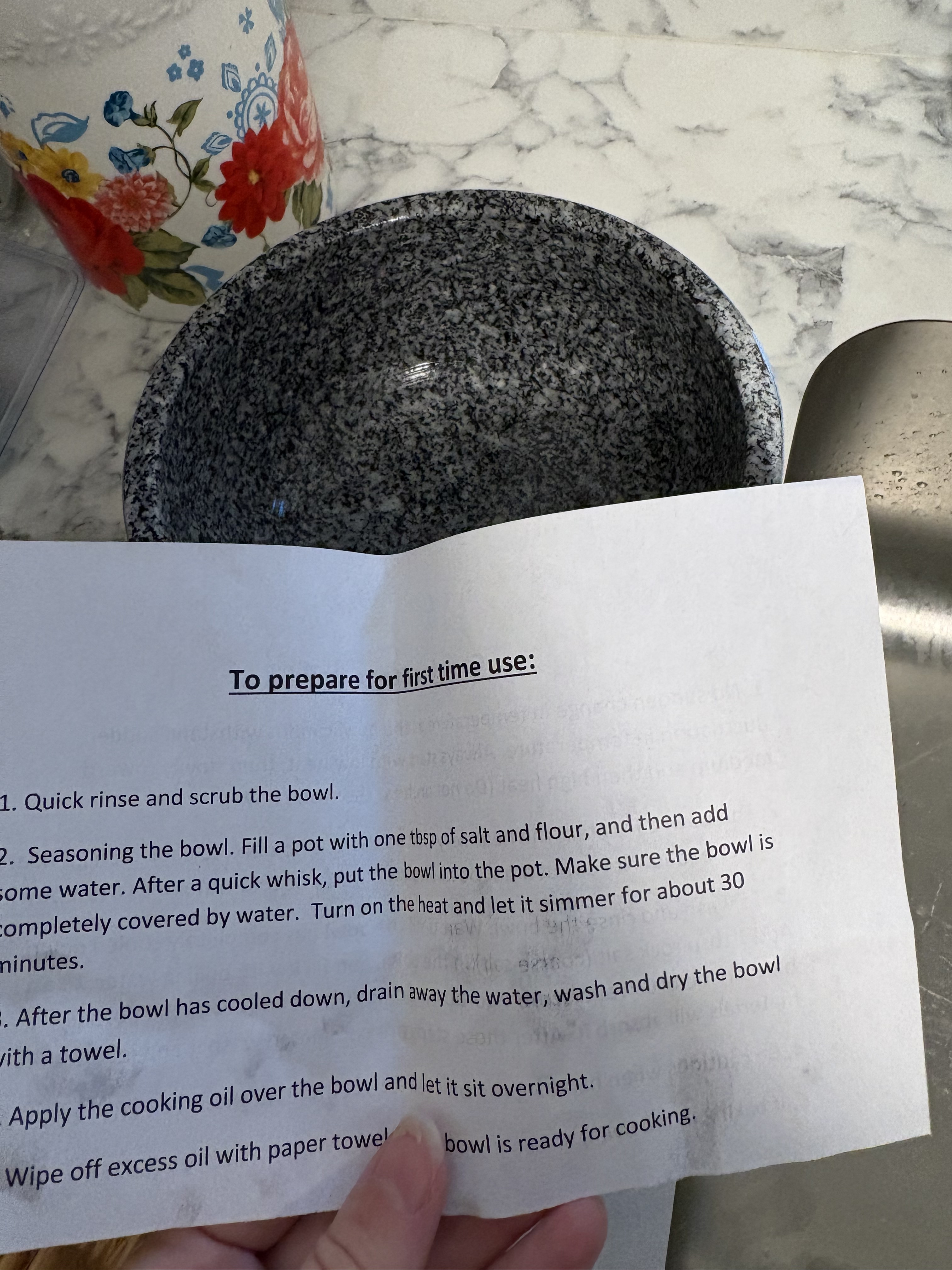 A first-use instruction leaflet is held in front of a speckled stone bowl; its seasoning step mentions salt and flour and a later step says to leave oil overnight.