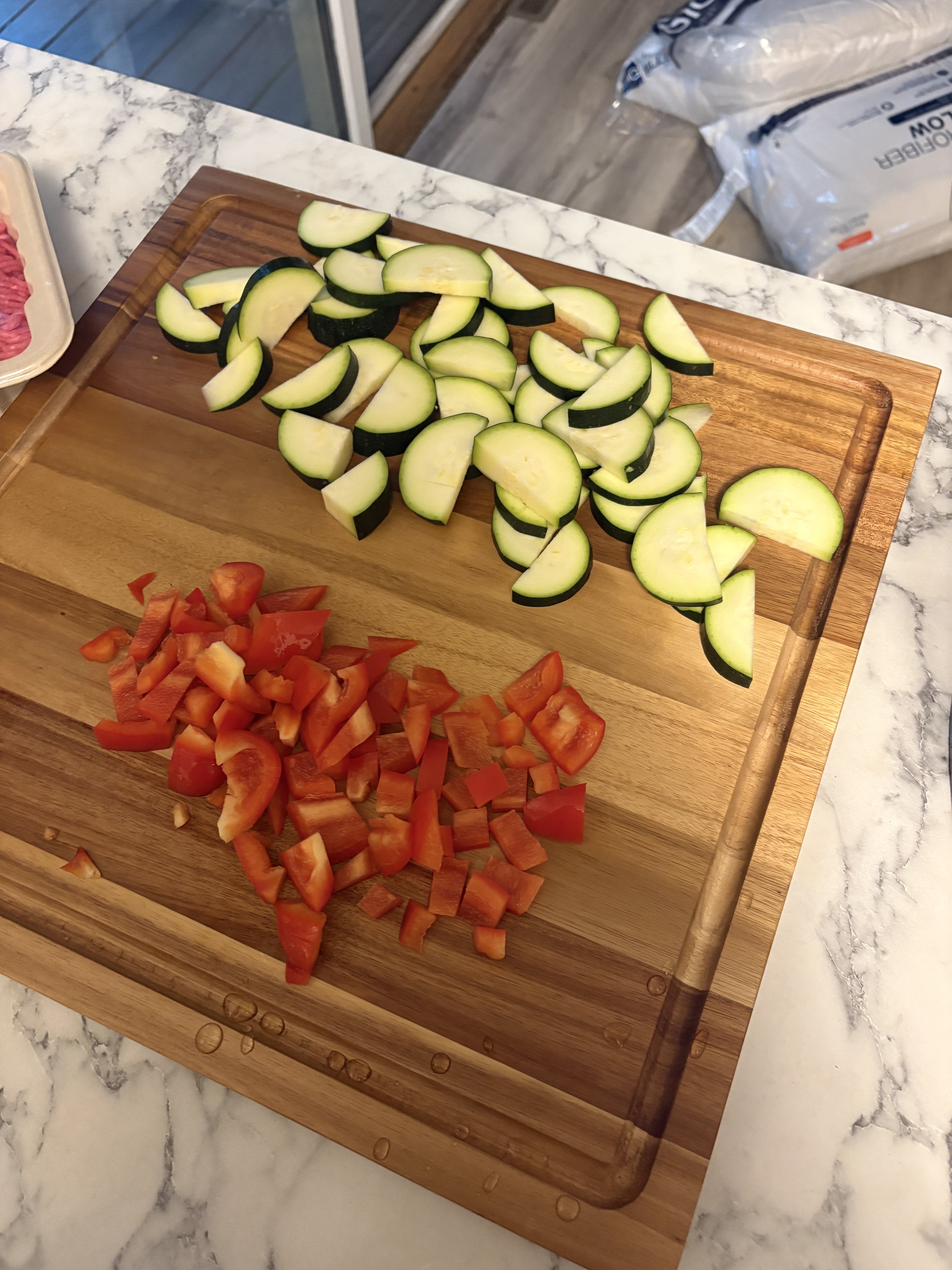 Sliced zucchini and chopped red bell pepper are arranged separately on a wooden cutting board.