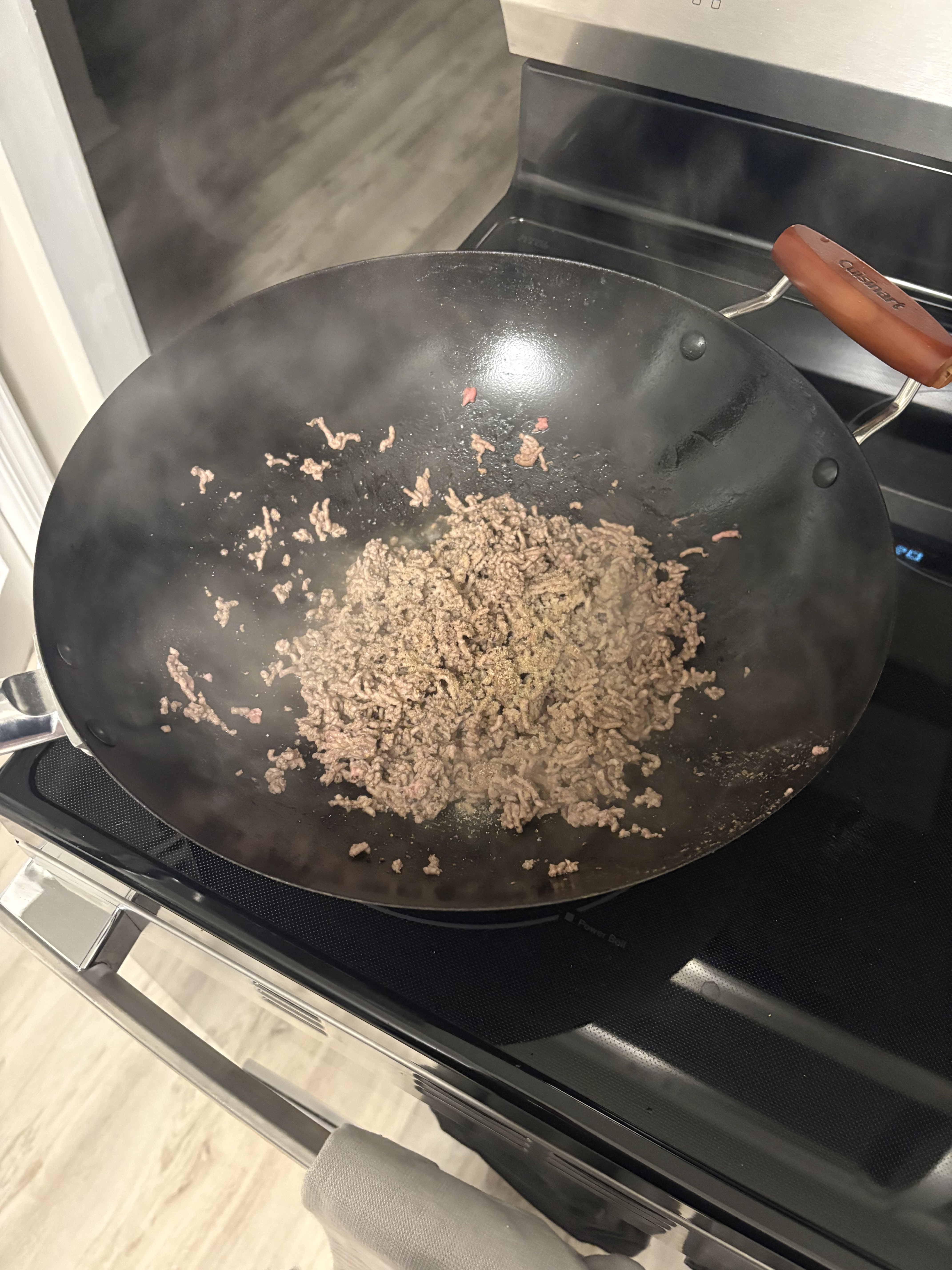 Ground beef is cooking in a black wok on a smooth cooktop, with steam visible above the pan.