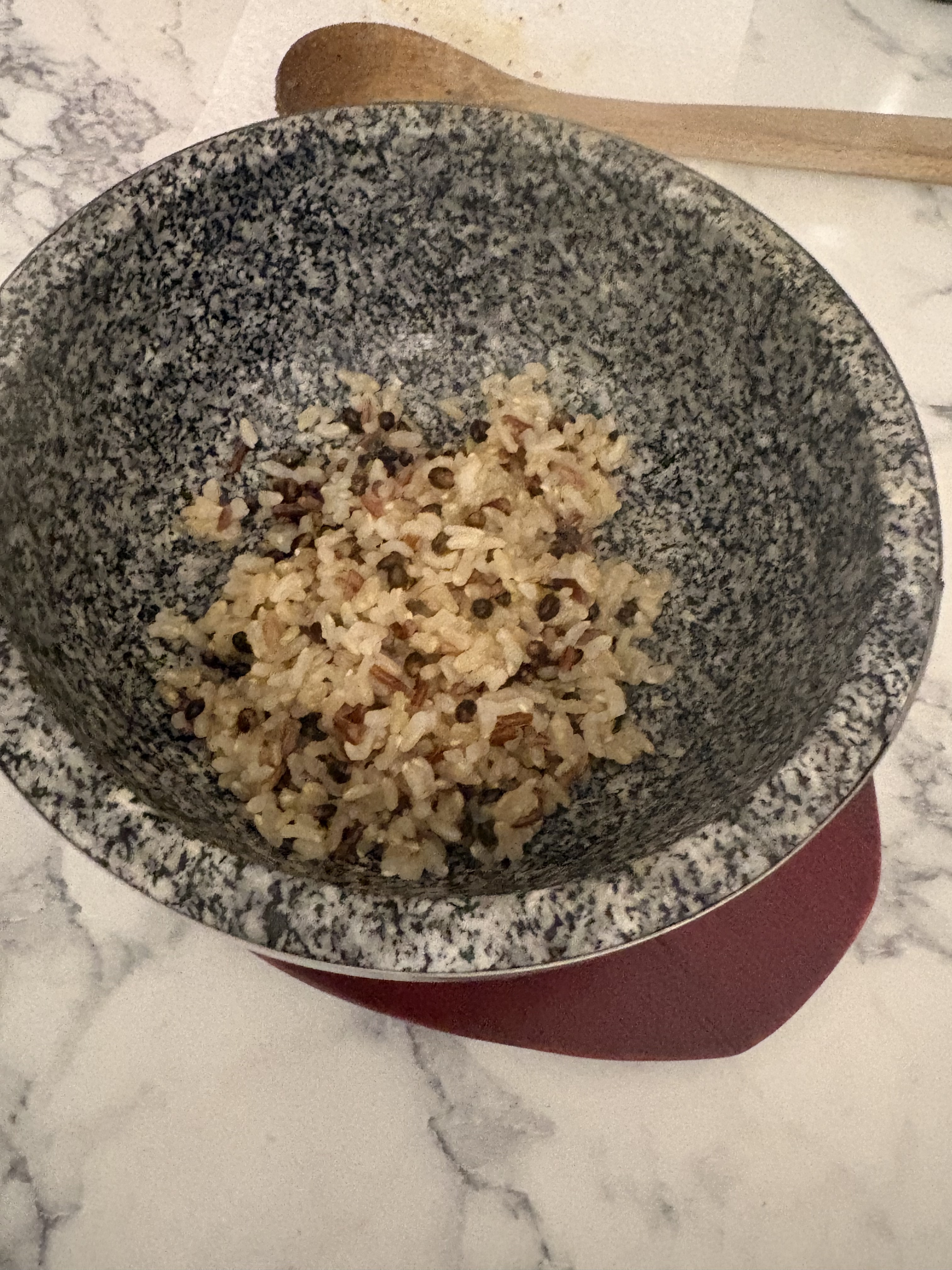 A portion of cooked rice with mixed colored grains sits in the bottom of a speckled stone bowl on a serving base.