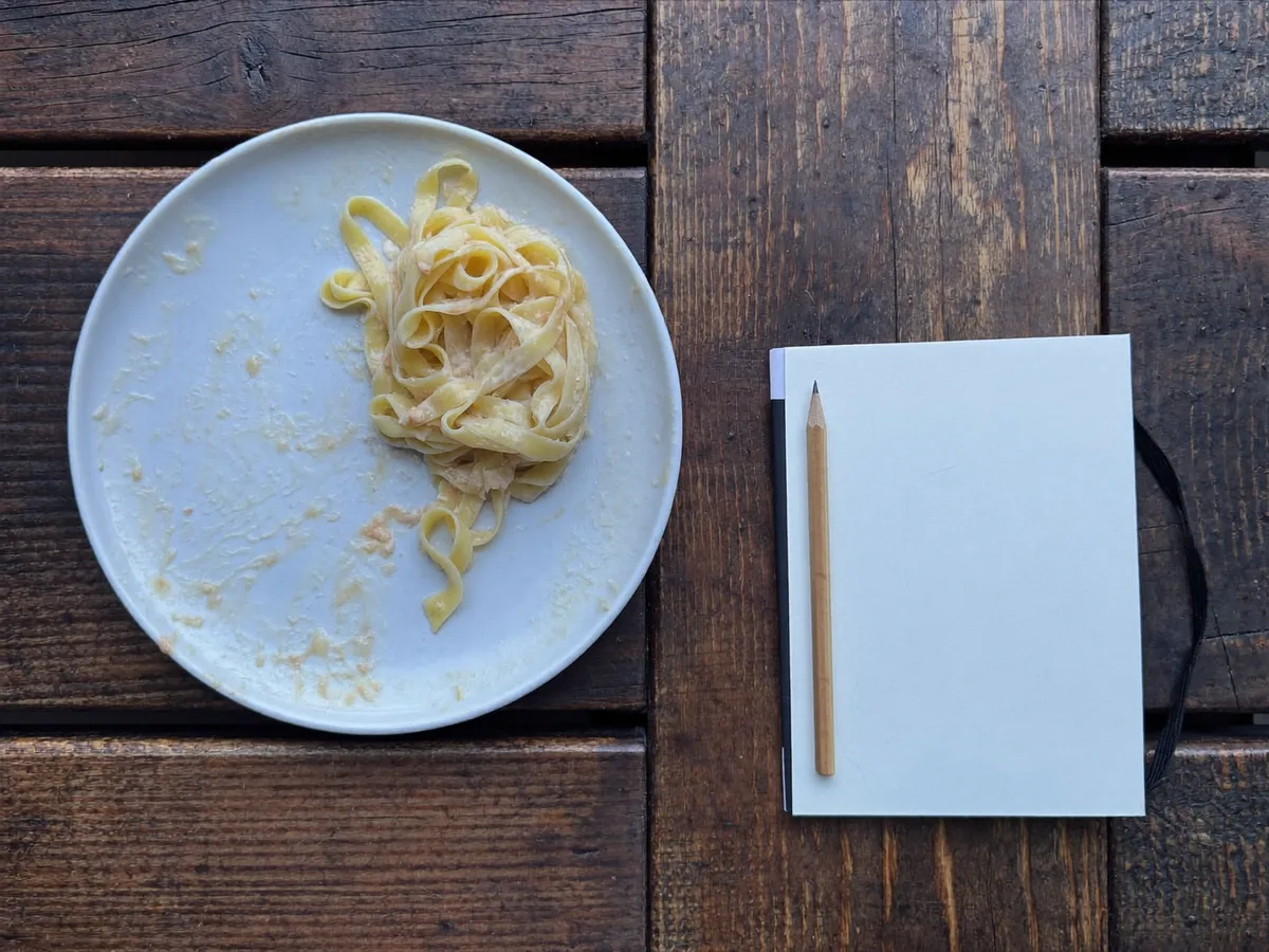 A blank notebook and pencil beside a plate of pasta